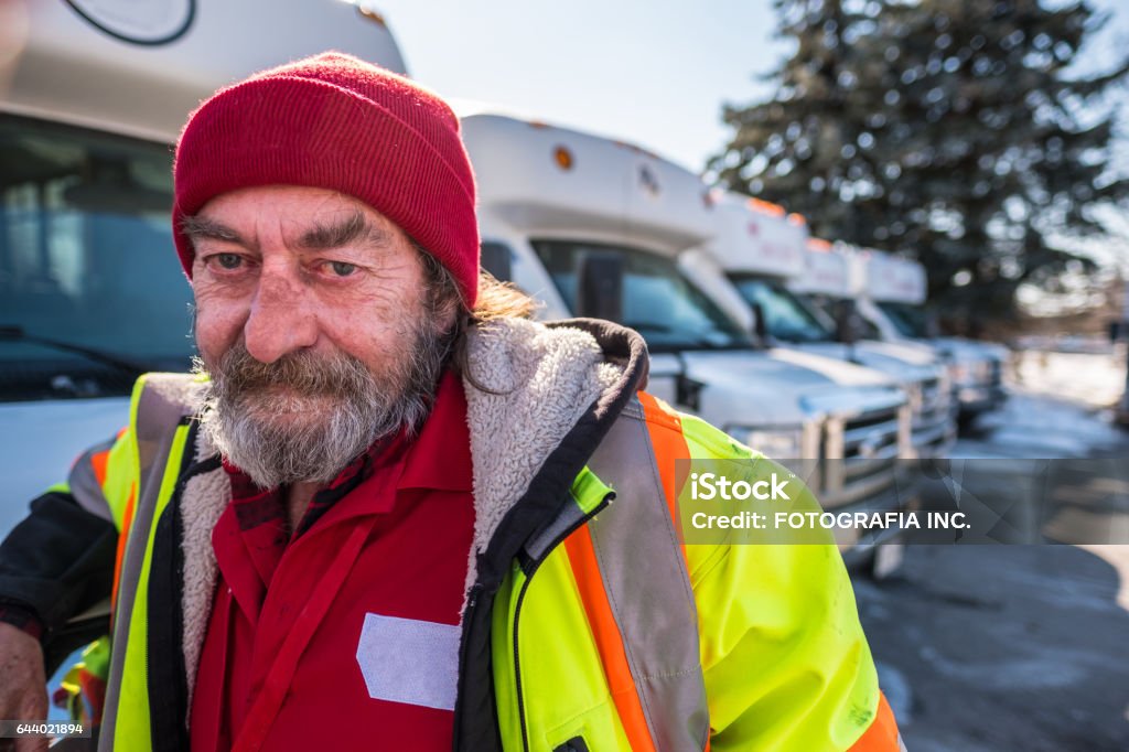 Winter Bus driver Location Portrait of male Bus driver standing outside row of parked  small size buses, looking at the camera. He is a mature man with gray haired beard. He is dressed in coat and hat. Bus Driver Stock Photo