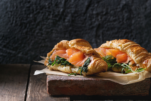 Two croissant with smoked salted salmon, spinach and arugula served on baking paper terracotta board over dark old wooden background. Copy space
