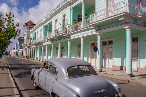 Photograph of a grey old classic car, driving on a street of downtown Cienfuegos. Next to the vehicle, a big aquamarine building, with columns and several balconies.