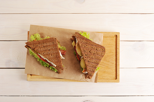two sandwiches from triangular pieces of bread with chicken breast, cheese and greens on paper on white wooden background - top view