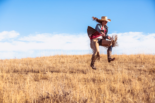 A silly young woman in cowboy boots, leather chaps and cowboy hat dancing on a grassy prairie.