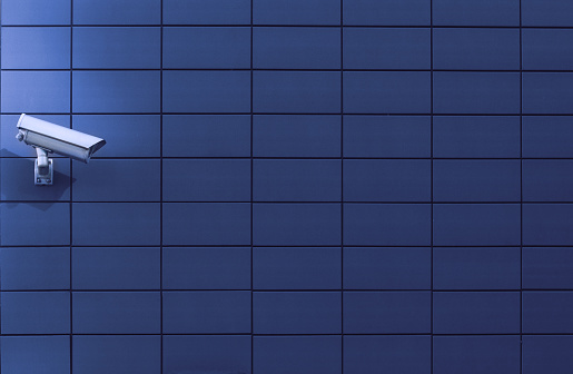 Horizontal front view of a surveillance monitoring white camera viewed from a side with blue background wall of metallic plates, copyspace on the right