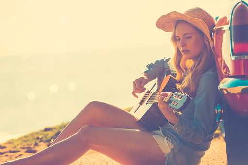 Woman with guitar leaning on a car. The car is parked at the beach at sunrise. Very relaxing vacation or road trip image. Car is a convertible. Focus on background. copy space