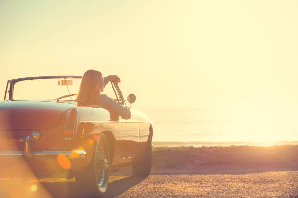 Young woman in a car at the beach. Young woman in a car at the beach. The car is a convertible, with the sunset and ocean backlit in the background. She looks relaxed and happy looking at the waterfront view. collectors car photos stock pictures, royalty-free photos & images