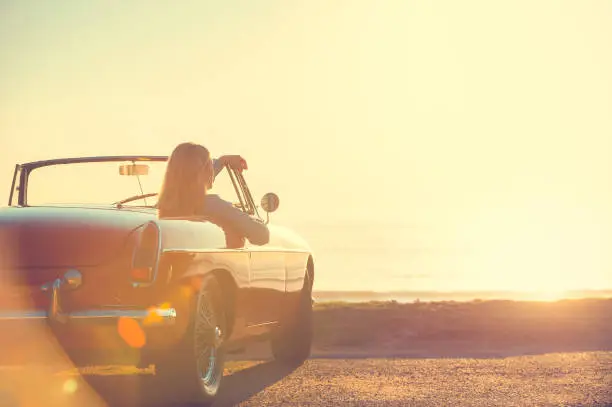 Young woman in a car at the beach. The car is a convertible, with the sunset and ocean backlit in the background. She looks relaxed and happy looking at the waterfront view.