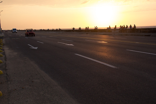 Havana, Cuba- March 26, 2016. Some old and classic cars driving on the  famous Cuban Malecon during a golden sunset. In the back, the silhouettes of some people seated watching the sunset and resting.