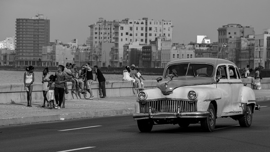 Havana, Cuba - March 26, 2016: Black and white photograph of a classic car driving on the boulevard next to the Malecon. There are some buildings in the bottom of the photograph, and several people sitted and walking on the side of the road.