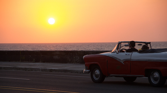 A couple driving, an old red and white classic car,  on the famous Cuban Malecon, during an orange  sunset
