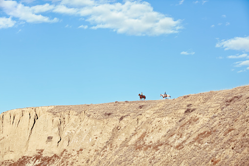 Two people on horseback, riding away on a distant high ridge under a blue sky with white clouds.