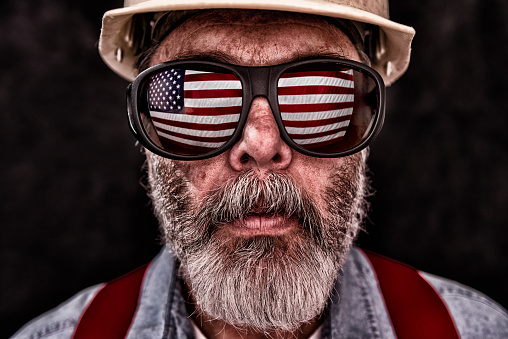 Close-up of grungy middle-aged Caucasian construction worker wearing eyeglasses with the reflection on the US flag in them. He is also wearing a worn white hard hat, blue denim shirt, red suspenders and a white t-shirt. He has an unkempt graying beard.