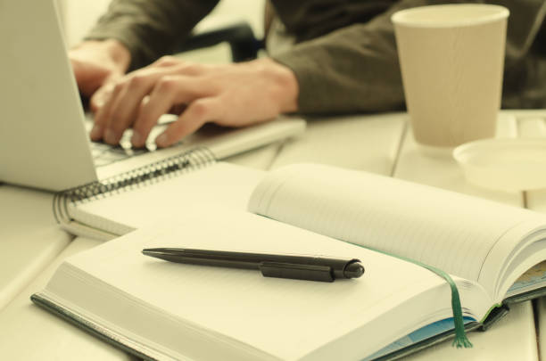 istock Open notepad with black pen on work-table. Paper cup of coffee, office stuff, laptop and working man on background 644144368