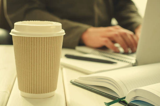 istock Paper cup of coffee on work-table with office stuff, notepad, laptop, pen, coffee cup and working man on background 644144374
