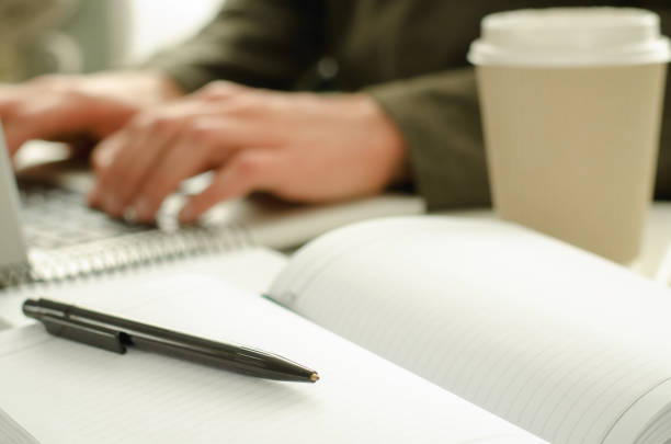 istock Open notepad with black pen on work-table. Paper cup of coffee, office stuff, laptop and working man on background 644144390