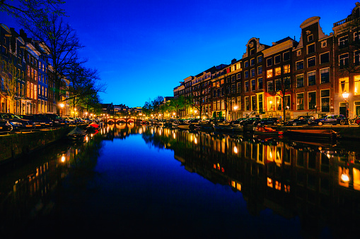Beautiful blue dusk cityscape in Amsterdam. Night illumination of Dutch buildings, parked boats and reflected city lights on canal water with blue sky.