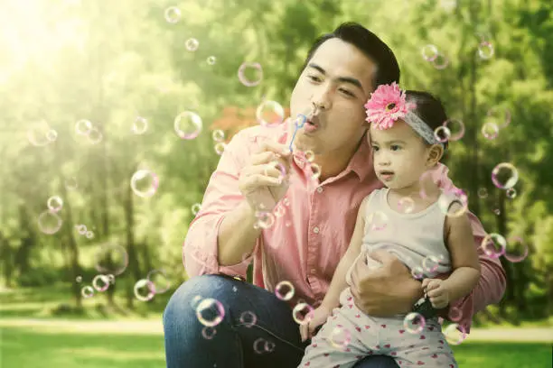 Image of young Asian father is blowing soap bubbles while embracing his daughter on the summer vacation