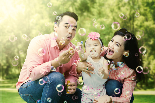 Image of young Asian parents blowing soap bubbles to their daughter while squatting at the playground
