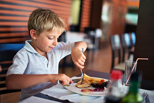 Little boy aged 7 having lunch with family in restaurant. The boy is focused on cutting the chicken meat with knife and fork.
