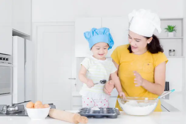Portrait of young mother and little daughter making cake together while sitting in the kitchen