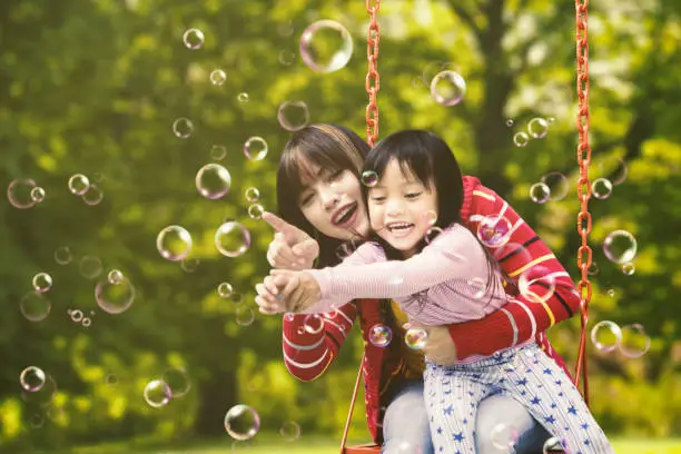 Pretty young mother and her little daughter sitting on a swing while touching soap bubbles at the park