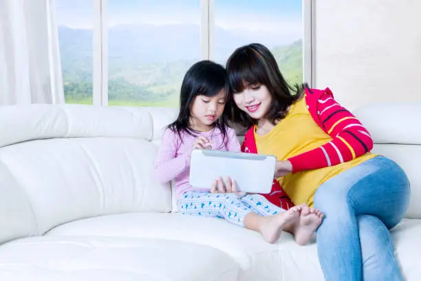 Photo of a pretty young woman and little girl using tablet while sitting on the couch at home