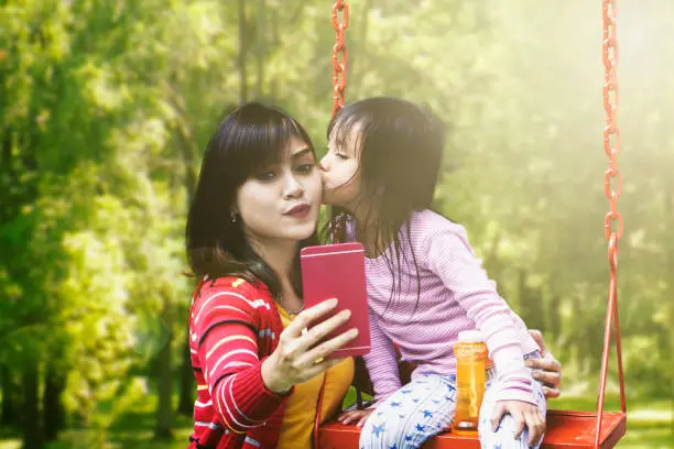 Happy young mother and her daughter taking selfie photo together while the girl kissing her mother at the park