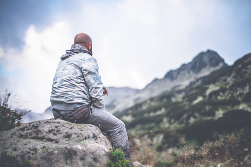 Male traveler sits on a rock alone in mountains and looking on a dramatic sky