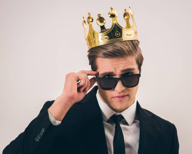 Handsome young prince Handsome young man in suit, crown and sun glasses is looking at camera while standing with crossed arms on gray background prince royal person photos stock pictures, royalty-free photos & images