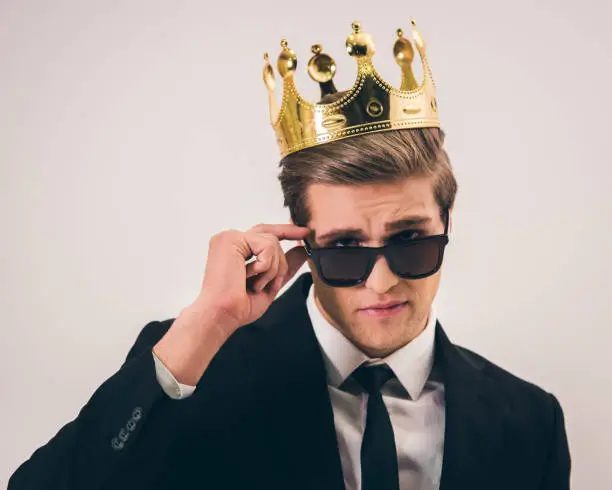 Handsome young man in suit, crown and sun glasses is looking at camera while standing with crossed arms on gray background