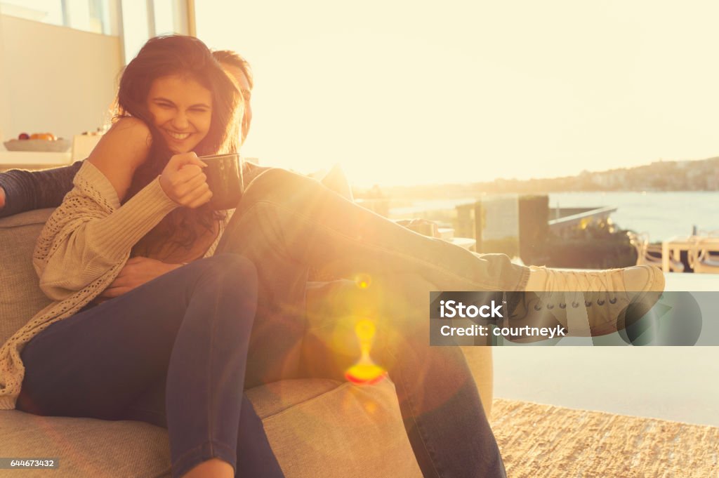 Couple on the sofa at sunrise. Couple on the sofa at sunrise. The woman has a cup of coffee. They are in a luxury house back lit by the sun with lens flare. Men Stock Photo