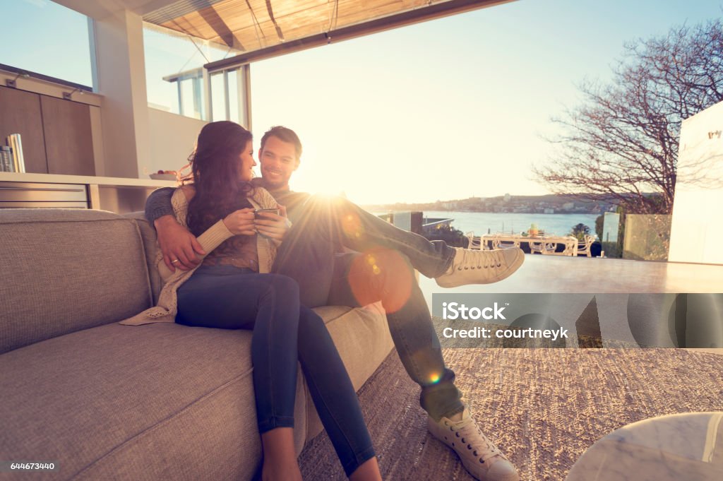 Couple on the sofa at sunrise. Couple on the sofa at sunrise. The woman has a cup of coffee. They are in a luxury house back lit by the sun with lens flare. Wide angle Luxury Stock Photo