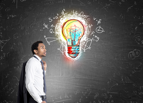 Side view of an African American man standing near a blackboard with a colorful light bulb surrounded by formulas.