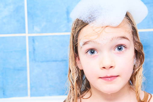 Adorable child blond girl with shampoo foam on hair taking bath. Closeup portrait of smiling kid, health care and hygiene concept.