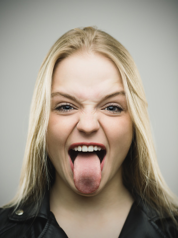 Close-up portrait of excited young woman with mouth open and tongue out. Cheerful female is grimacing. She is against gray background. Vertical studio photography from a DSLR camera. Sharp focus on eyes.