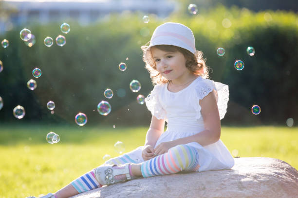 Adorable little girl, sitting on a rock, surrounded with soap bubbles, summer sunbeams, dreaming. Carefree child on green summer meadow. Happiness, childhood and freedom concept.