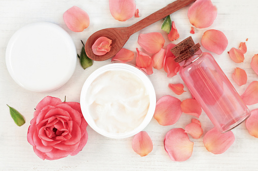with pink rose petals. Jar of body moisturizer, attar bottle toning lotion, top view homemade cosmetic ingredients.