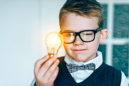 Boy with glasses holds a light bulb in his hand. It lights up: concept of getting a good idea. The boy also wears a bow tie.