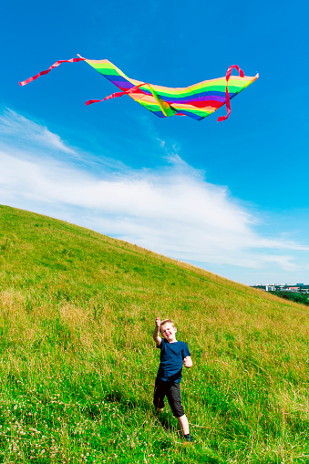 Boy stands on a hill and holds on to a string attached to a kite. The kite is multi colored and flys high in the air. It is summer.