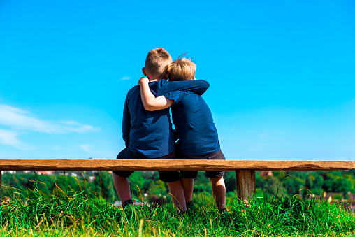 Two friends or brothers sit on a bench outside. They sit close and hug each other.