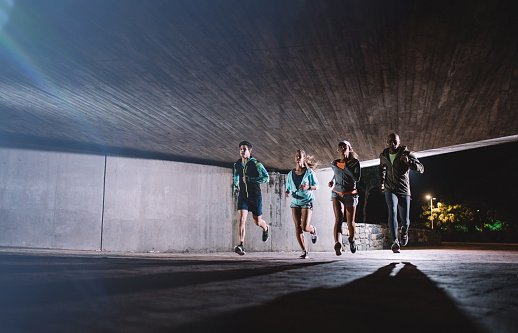 Group of young men and women running together at night. Healthy young people training together under a bridge in city.