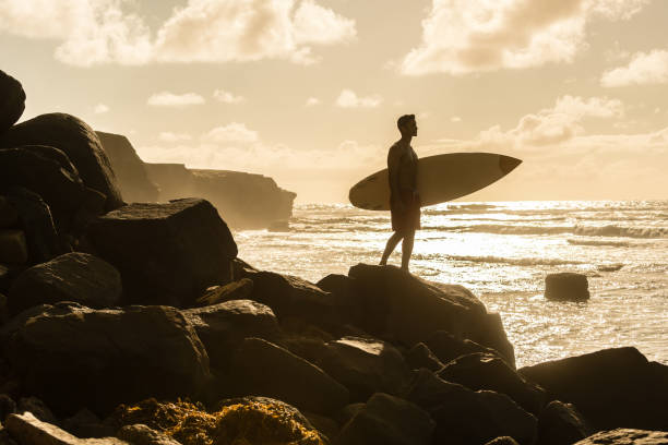 A hispanic man posing with his surfboard with a rugged coast line behind.