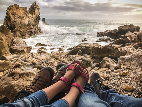 Family, including a mother, father and daughter rest on the shores of a rocky beach together. Shot on the beach of Corona del Mar in Orange County, Southern California.  Shot from their point-of-view, showing their legs and feet. They're wearing protective shoes.