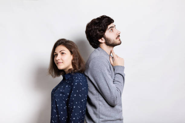 istock A horizontal portrait of two young people standing back to back having thoughtful expression. A stylish young bearded man holding his finger on his neck looking up thinking standing back to his girl 645243114