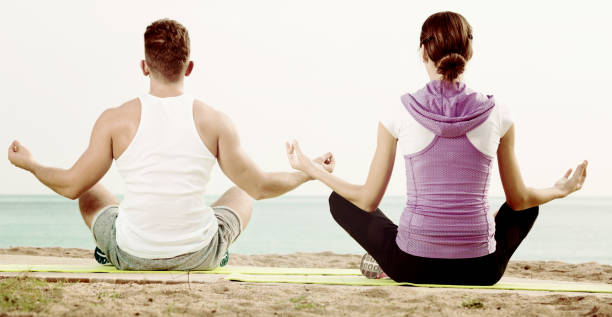 mulher e homem sentado de pernas cruzadas fazem poses de ioga na praia - men yoga meditating cross legged - fotografias e filmes do acervo