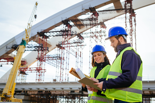 Two engineers, male and female, in protective helmets and reflective clothing, standing on construction site where large arch bridge is being built. Engineers looking at blueprints, planning and discussing project development. Image taken in Central Europe, bridge is on Danube river