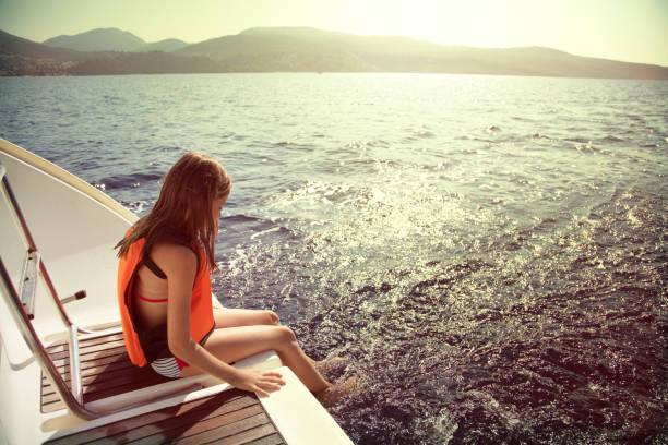 Little girl sitting on the  back part of the ship wihile sailing on the Ionian sea in Greece. She put their feet in the blue sea and enjoy while waves massaging it. Girl is wearing a life jackets.