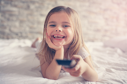 Little girl watching TV lying on bed with remote control in hand.