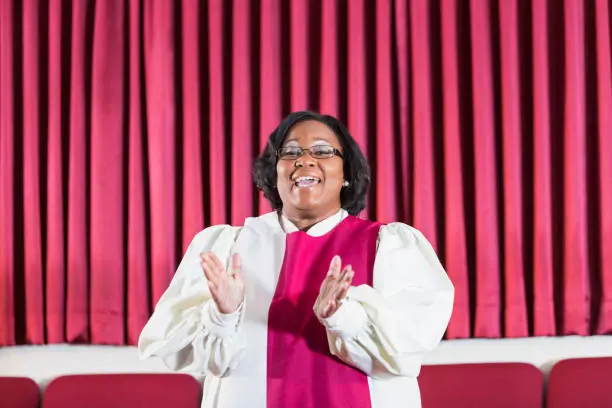 A mature black woman singing in a church choir. She is singing and clapping.