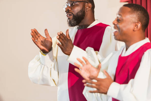 Mature black men singing gospel music in church choir Two mature black men singing gospel music in a church choir. They are wearing robes, singing and clapping. The focus is on the man with a beard wearing eyeglasses. gospel stock pictures, royalty-free photos & images