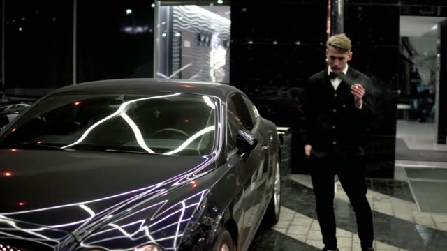 Portrait of a young man in a black suit with a bow-tie smoking a cigarette by the black luxury car at night.