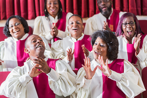 A group of mature African American women and men wearing robes, singing and clapping in a church choir.